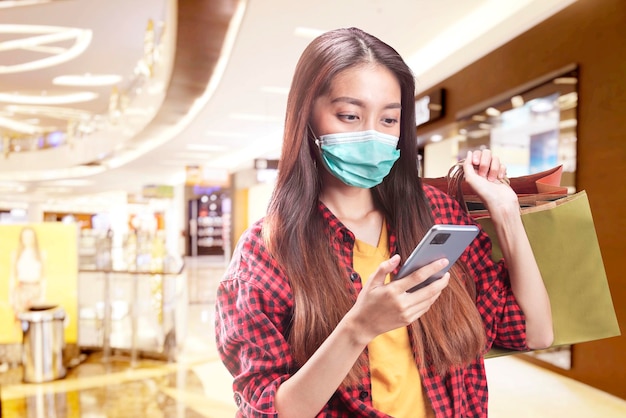Femme asiatique en masque facial tenant un téléphone portable tout en portant des sacs à provisions dans le centre commercial