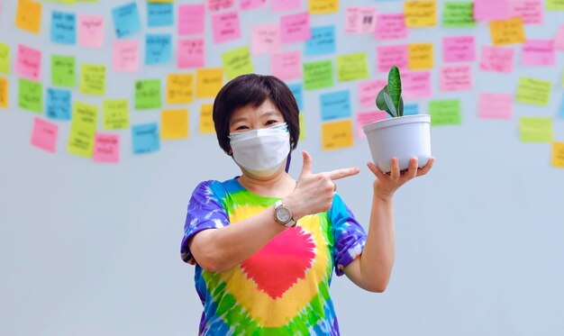 Femme asiatique en masque de protection montrant une plante d'intérieur avec un arrière-plan flou de notes autocollantes sur un mur gris