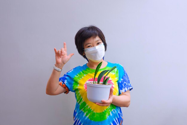 Femme asiatique en masque de protection montrant sa plante d'intérieur et signe de la main d'amour sur le mur de ciment gris