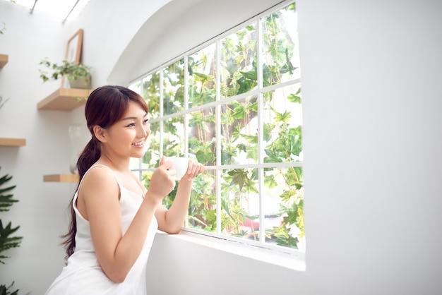 Femme asiatique le matin tenant une tasse de thé ou de café et debout derrière la fenêtre