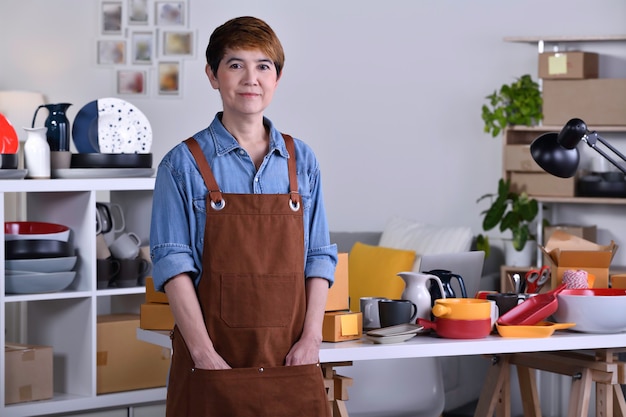 Femme asiatique mature entrepreneur/ propriétaire d'entreprise debout devant son produit en céramique d'argile et travaillant à la maison