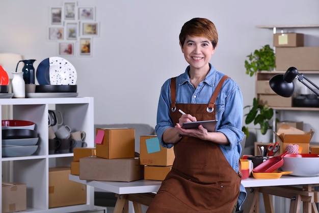 Femme asiatique mature entrepreneur/ propriétaire d'entreprise debout devant son produit en céramique d'argile et travaillant à la maison