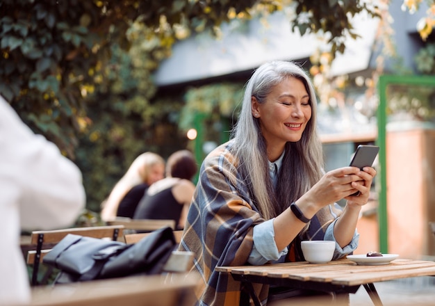 Une femme asiatique mature et heureuse surfe sur Internet au téléphone à une petite table sur la terrasse du café en plein air