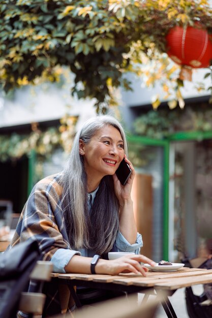 Une femme asiatique mature et positive parle sur un téléphone portable à une petite table dans un café en plein air