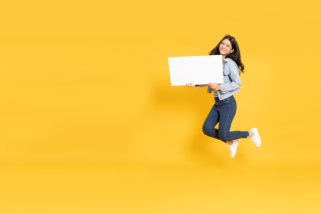 Femme asiatique montrant et tenant un panneau d'affichage blanc vierge et sautant isolé sur fond jaune