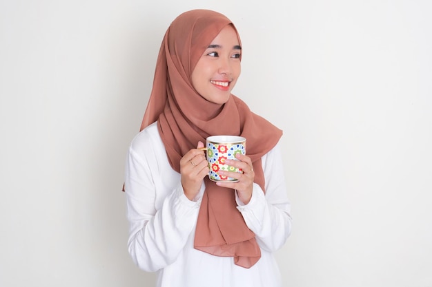 Une femme asiatique musulmane détournant le regard tout en tenant un verre à boire avec une expression heureuse