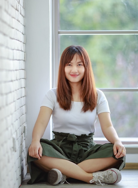 Femme asiatique paisible assise sur le rebord de la fenêtre contre la fenêtre et regardant la caméra à la maison