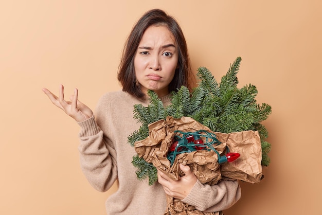 Une femme asiatique perplexe et indignée hausse les épaules a des regards déplus à l'expression avec perplexité tient une guirlande du Nouvel An et des branches de sapin enveloppées dans du papier isolé sur fond beige.