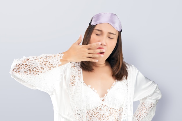 Femme asiatique portant une chemise de nuit en dentelle blanche et une robe en satin bâillant montrant un geste endormi couvrant la bouche avec sa main.