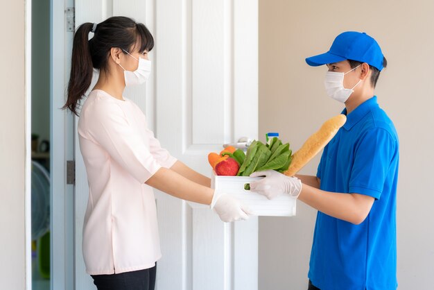 Une femme asiatique portant un masque facial et un gant reçoit une boîte d'épicerie de nourriture, de fruits, de légumes et de boissons d'un livreur devant la maison pendant l'isolement.