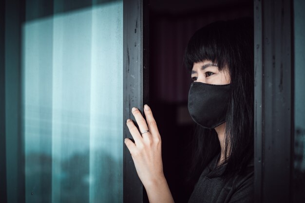 Femme asiatique portant un masque noir, distrait à la porte et restant à la maison pour l'auto-quarantaine et la distanciation sociale dans le coronavirus ou Covid-2019 concept de situation d'épidémie