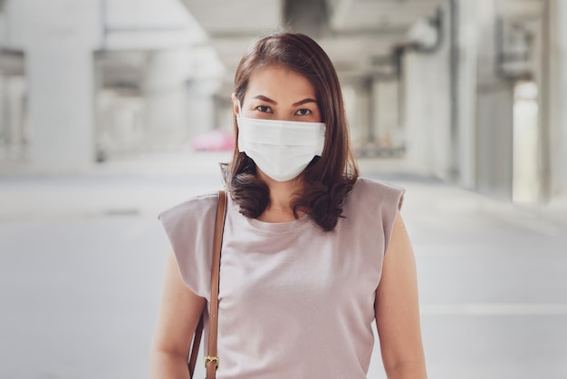 Femme asiatique portant un masque de protection debout et regardant la caméra à l'extérieur dans un contexte de construction urbaine avec espace de copie. Concept de nouveau mode de vie normal et empêche le virus COVID-19.