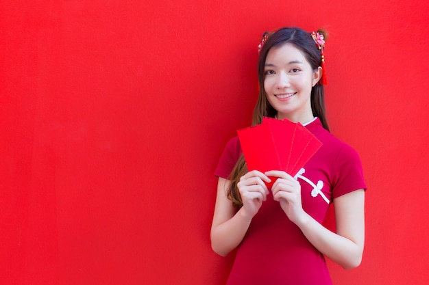 Une femme asiatique porte un cheongsam rouge et tient des enveloppes rouges tout en regardant la caméra et en souriant.