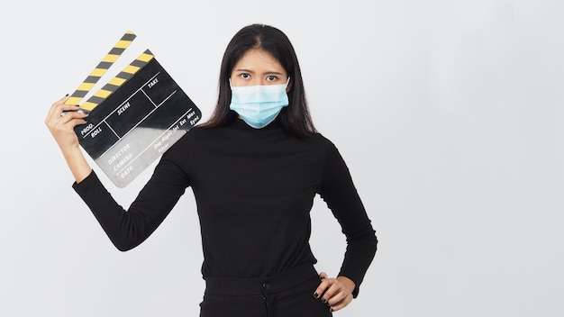 Une femme asiatique porte un masque facial et une main tenant un panneau de clapet noir ou une ardoise de film dans la production vidéo, le cinéma, l'industrie du cinéma sur fond blanc.