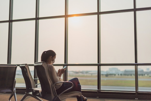 Une femme asiatique porte un masque facial et utilise un smartphone dans l'arrière-plan du terminal de l'aéroport. Concept de transport public Soigneusement pendant l'épidémie de COVID 19. et de coronavirus, distanciation sociale.