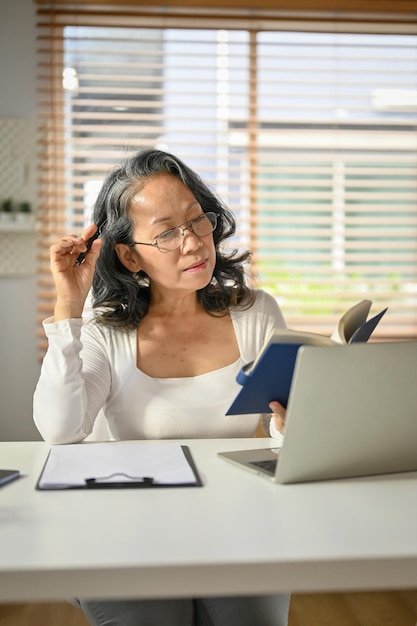 Femme asiatique positive des années 60 se concentrant sur la lecture d'un livre dans son espace de travail