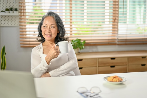 Femme asiatique positive regardant par la fenêtre rêvant d'avoir son café du matin