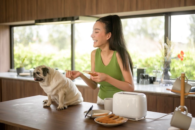 Une femme asiatique prépare du café et du pain grillé pour le petit déjeuner avec un chien dans la cuisine.