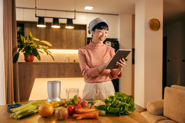 Une femme asiatique prépare un smoothie sain à la maison en souriant à la tablette et en faisant défiler sur elle