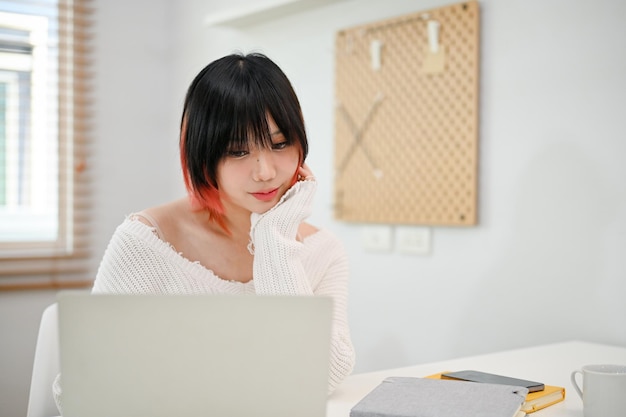 Femme asiatique réfléchie se concentrant sur ses tâches sur un ordinateur portable naviguant sur Internet