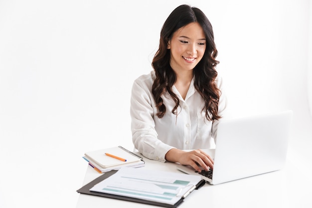 Femme asiatique réussie avec de longs cheveux noirs assis à table et travaillant avec des documents et un ordinateur portable