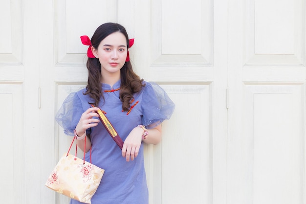 Une femme asiatique en robe qipao chinoise bleu-gris tient le sac en tissu sur une porte en bois blanc en arrière-plan