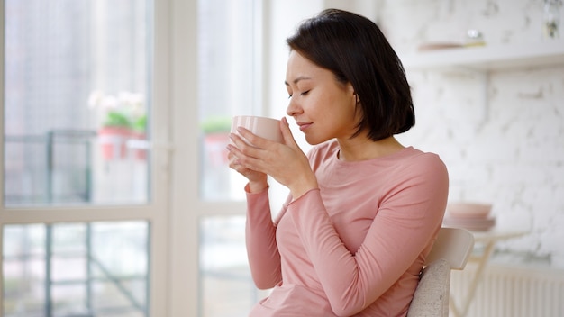 Femme asiatique satisfaite multiethnique appréciant une tasse de café ou de thé buvant une boisson chaude à la maison