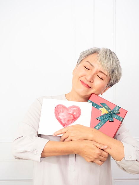 Femme asiatique senior heureuse souriante et tenant une boîte-cadeau et une carte de voeux dans les mains.