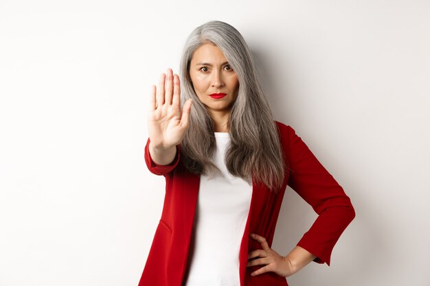 Femme asiatique sérieuse en blazer rouge montrant un panneau d'arrêt, disant non, fronçant les sourcils et rejetant quelque chose de mal, debout sur fond blanc