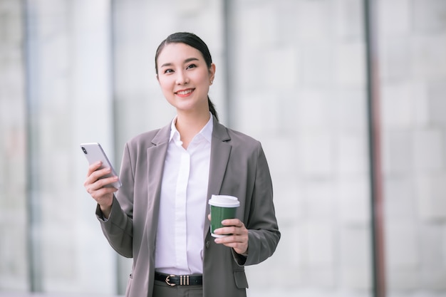 Femme asiatique avec smartphone debout contre le bâtiment flou de la rue. Photo d'entreprise de mode de belle fille dans une suite décontractée avec téléphone et tasse de café.