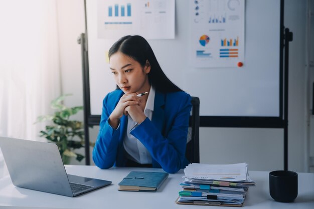 Une femme asiatique souffrant de migraines, de tensions cérébrales, d'une femme d'affaires surchargée de travail, d'un financier travaillant sur un ordinateur portable et une tablette dans un bureau moderne.