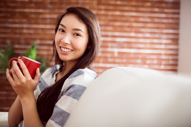 Femme asiatique souriante sur le canapé ayant une boisson chaude