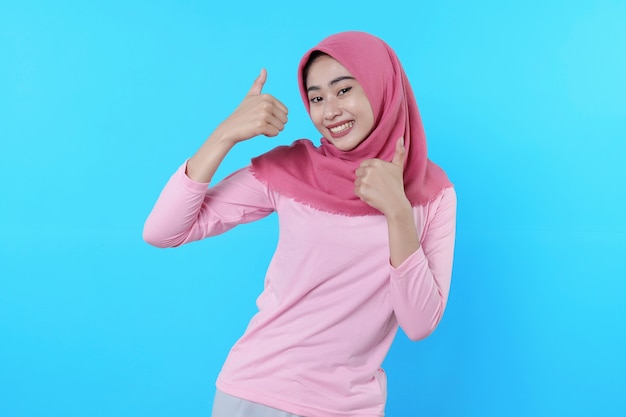 Femme asiatique souriante avec un coup de pouce isolé sur fond bleu clair portant un hijab avec un t-shirt rose