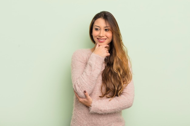 Femme asiatique souriante avec une expression confiante heureuse avec la main sur le menton se demandant et regardant sur le côté