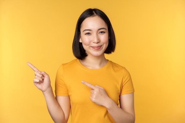 Femme asiatique souriante pointant les doigts vers la gauche montrant une publicité sur un espace de copie vide debout sur fond jaune