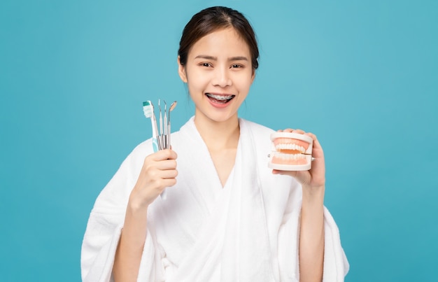 Femme asiatique souriante portant des bretelles avec une dent et tenant une brosse à dents et un outil de dentiste isolé sur fond bleu, Concept d'hygiène buccale et de soins de santé.