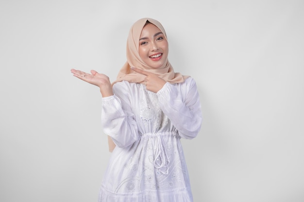 Une femme asiatique souriante portant un hijab et une robe blanche pointant vers l'espace de copie sur son côté droit debout sur un fond blanc isolé concept publicitaire Ramadan et Eid Mubarak