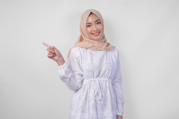 Une femme asiatique souriante portant un hijab et une robe blanche pointant vers l'espace de copie sur son côté droit debout sur un fond blanc isolé concept publicitaire Ramadan et Eid Mubarak