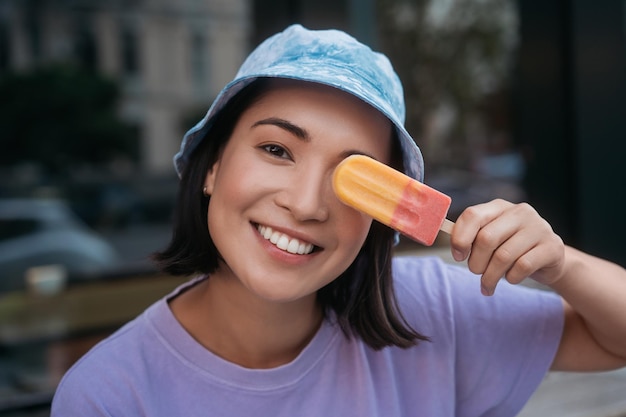 Femme asiatique souriante portant un panama élégant tenant une glace près du visage en regardant la caméra