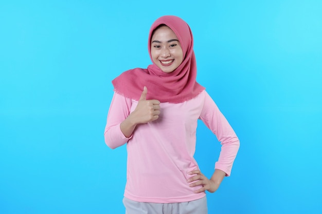 Femme asiatique souriante avec un pouce isolé sur fond bleu clair portant un hijab avec un t-shirt rose