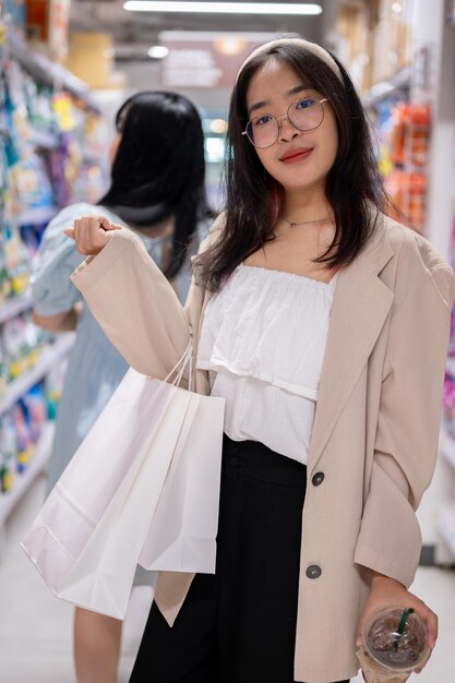 Une femme asiatique souriante se tient dans un couloir d'épicerie tenant des sacs d'achat dans un supermarché