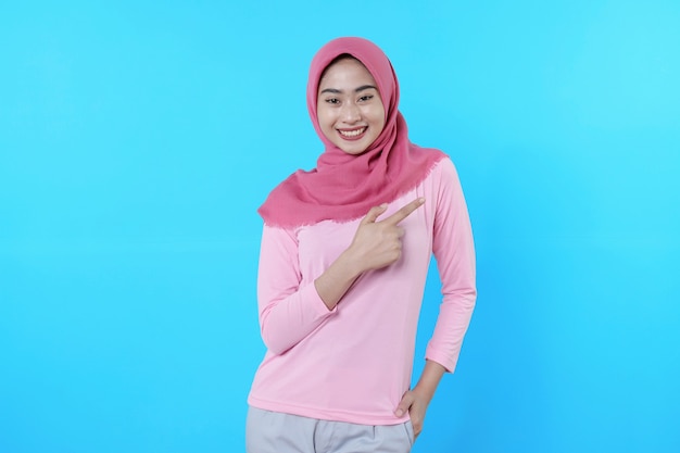 Femme asiatique souriante avec son doigt pointé isolé sur fond bleu clair portant un hijab avec un t-shirt rose