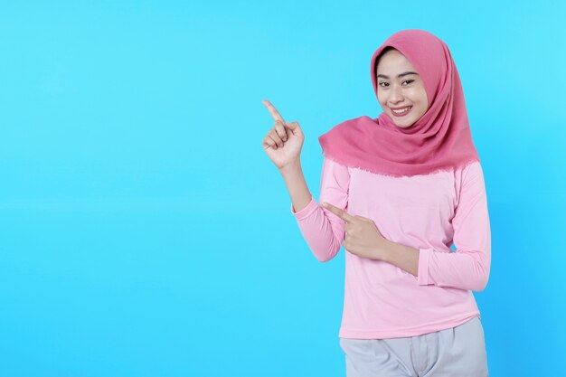 Femme asiatique souriante avec son doigt pointé isolé sur fond bleu clair portant un hijab avec un t-shirt rose