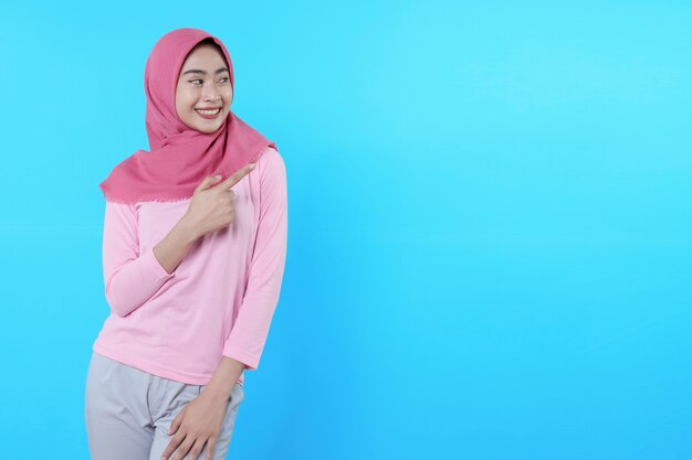 Femme asiatique souriante avec son doigt pointé isolé sur fond bleu clair portant un hijab avec un t-shirt rose