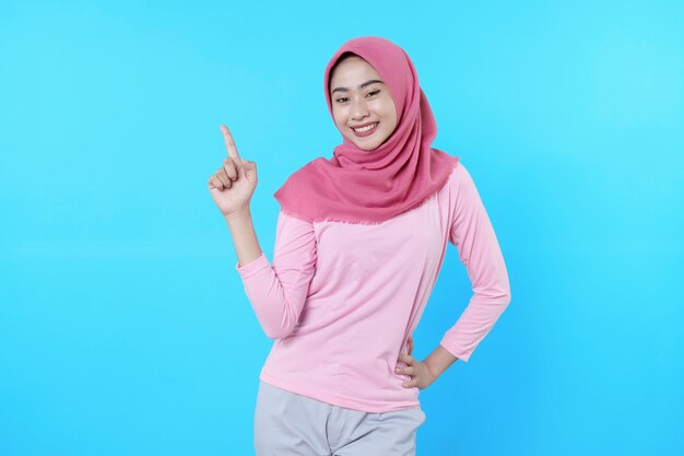 Femme asiatique souriante avec son doigt pointé isolé sur fond bleu clair portant un hijab avec un t-shirt rose