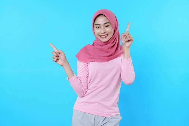 Femme asiatique souriante avec son doigt pointé isolé sur fond bleu clair portant un hijab avec un t-shirt rose