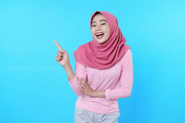 Femme asiatique souriante avec son doigt pointé isolé sur fond bleu clair portant un hijab avec un t-shirt rose