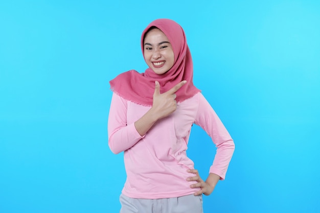 Femme asiatique souriante avec son doigt pointé isolé sur fond bleu clair portant un hijab avec un t-shirt rose