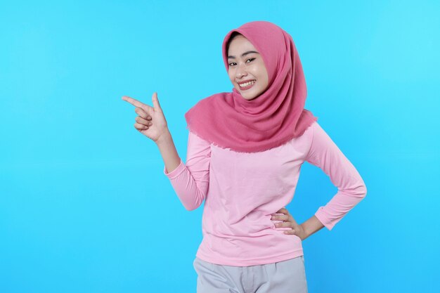 Femme asiatique souriante avec son doigt pointé isolé sur fond bleu clair portant un hijab avec un t-shirt rose