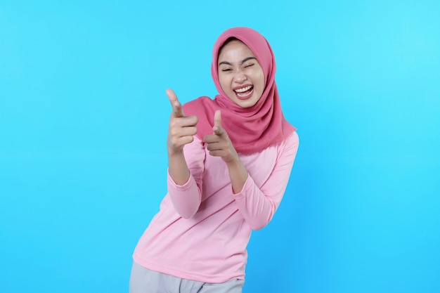 Femme asiatique souriante avec son doigt pointé isolé sur fond bleu clair portant un hijab avec un t-shirt rose